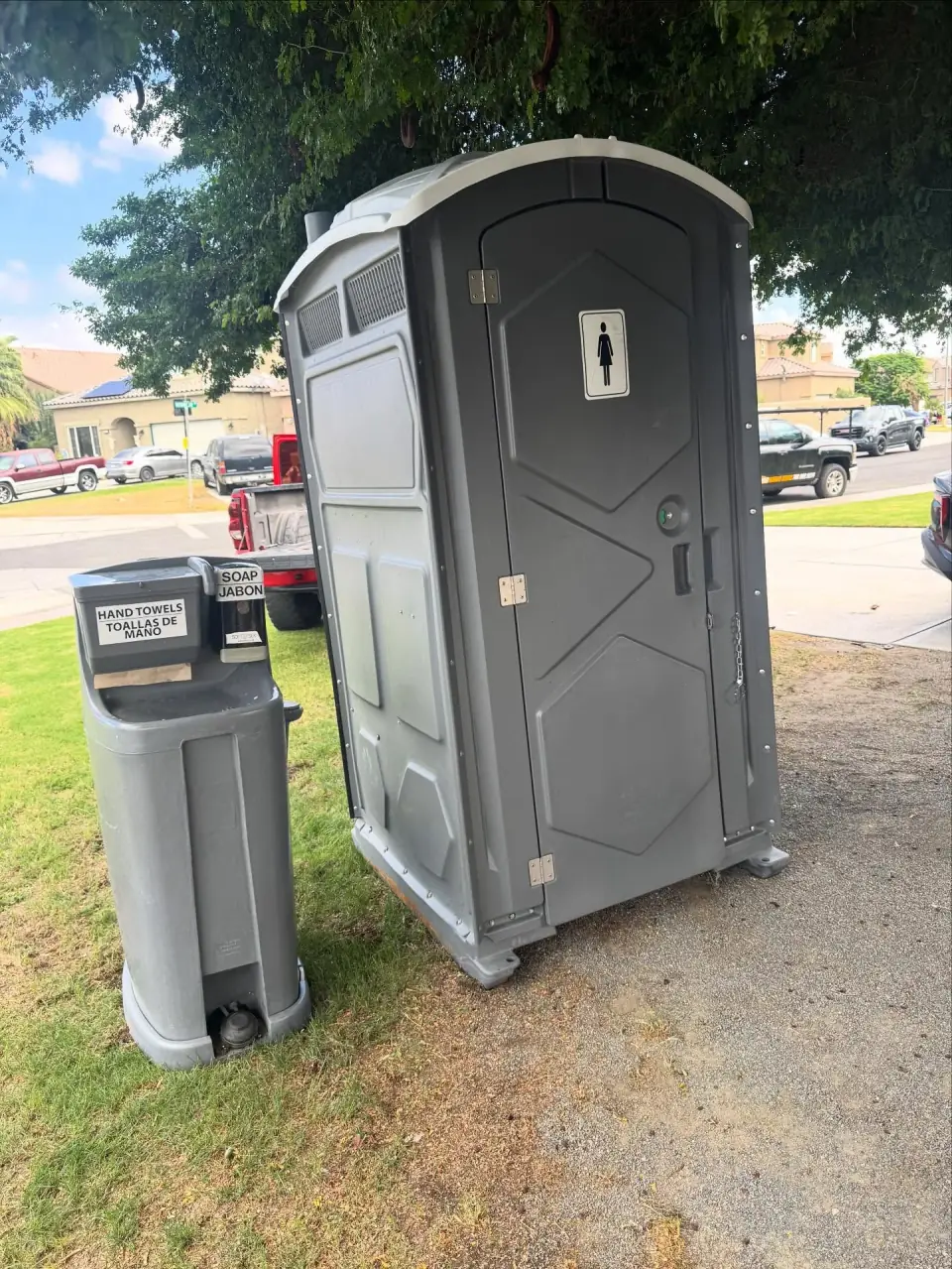 Freestanding gray portable toilet beside a handwashing station on a lawn